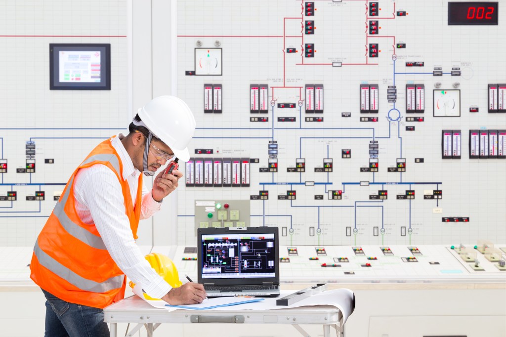 An electrical engineer in a hard hat and orange vest works in a control room