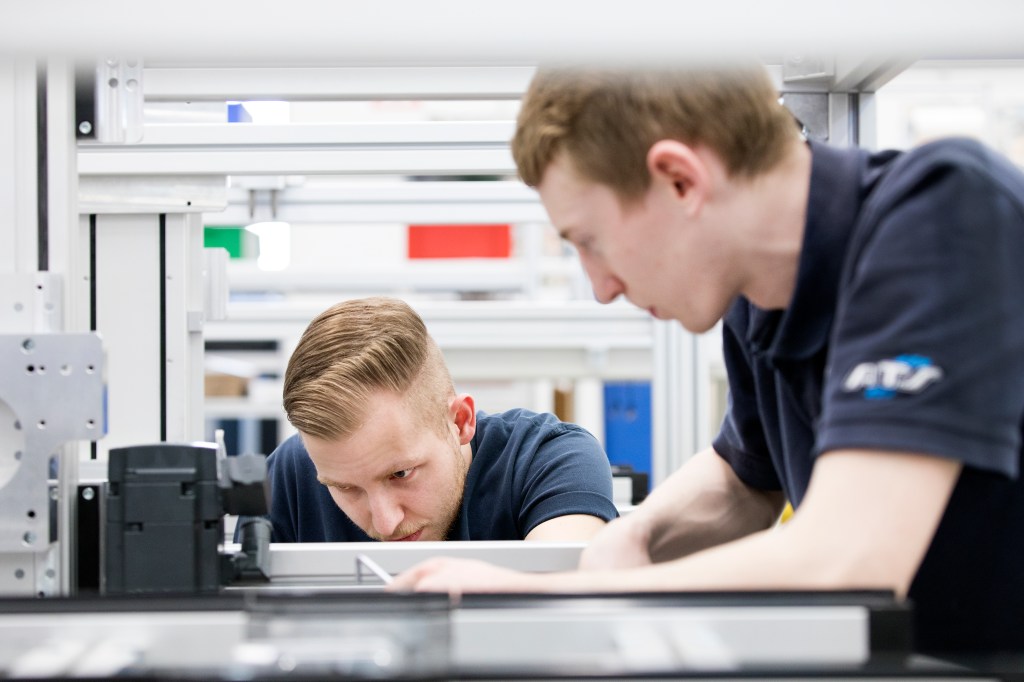 A male employee demonstrates for another male employee how to assemble part of a machine