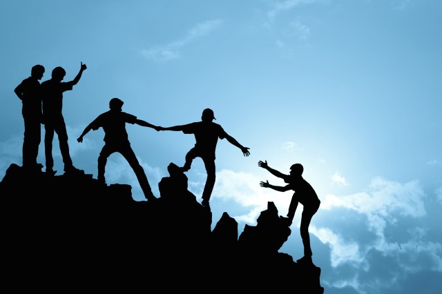 Immagine della silhouette di un team di persone, che si aiutano tra loro a salire in cima a un piano inclinato su uno sfondo con cielo azzurro